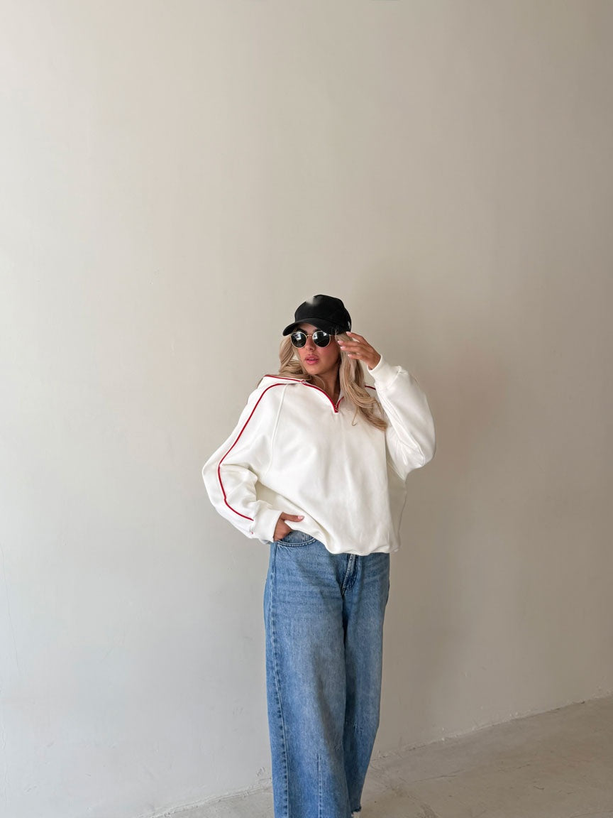 Femme en sweat demi-zip blanc à passepoil rouge, main sur la casquette